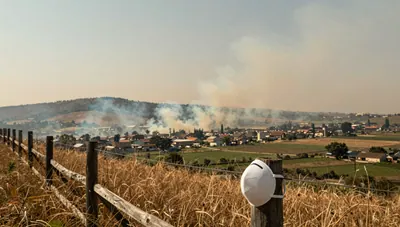 Smoke plume from a distant hillside drifting over a hazy valley town, with a mask on a fence post in the foreground.