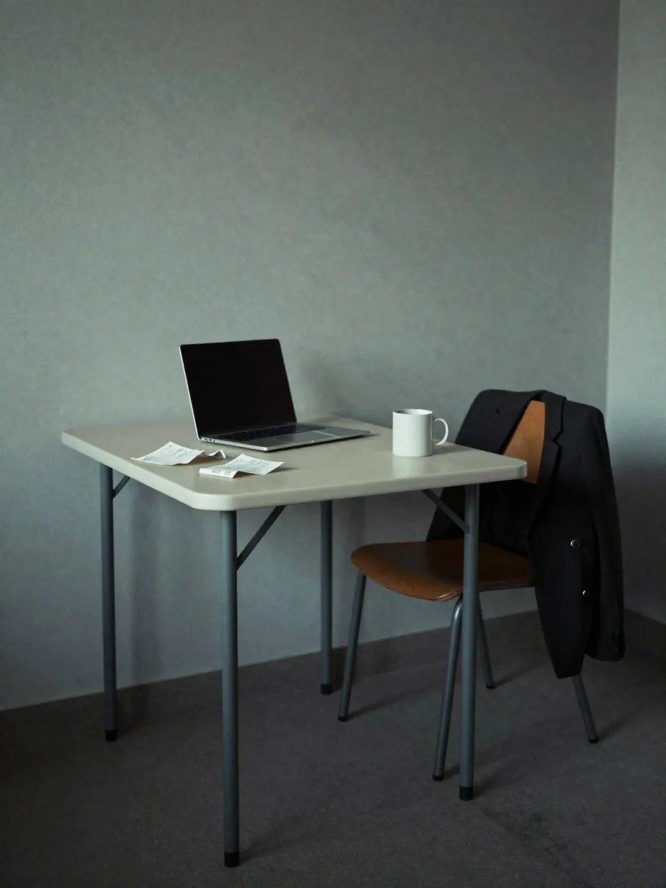 Sparse rented flat with a folding table, laptop, receipts, mug and a coat over a chair.