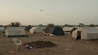 Dusk over a dusty camp of patched tents and tarpaulins, cooking fires glowing, a small aircraft silhouette high in a hazy sky.