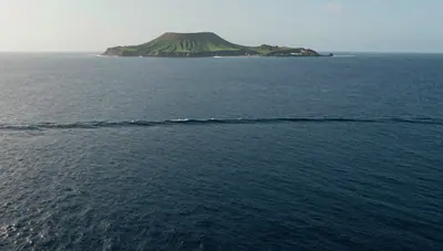 Aerial view of a long, low tsunami swell crossing deep open ocean toward a distant volcanic island.