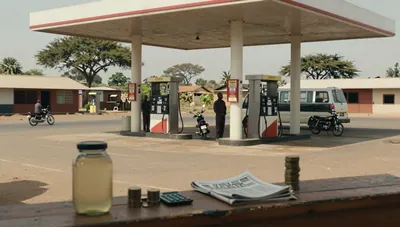 Roadside Kenyan fuel station with pumps, attendant, matatu and motorcycle, coins and a jar of diesel on a counter in the foreground.