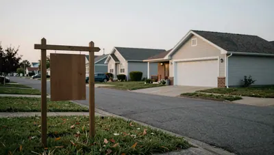 Empty real-estate sign on a quiet suburban lawn, closed houses across the street, overcast afternoon light.