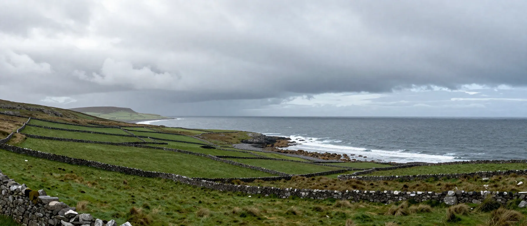 Panoramic view of green Irish hills and rocky coast beneath rain bands and broken Atlantic cloud.