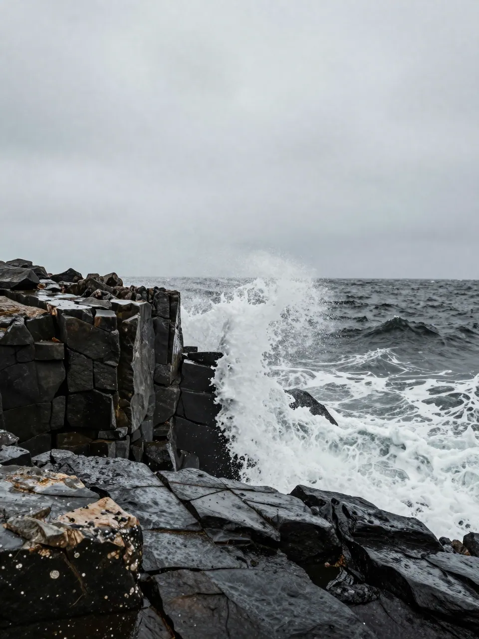A dark wave breaking hard against a rocky coastline under heavy overcast sky.