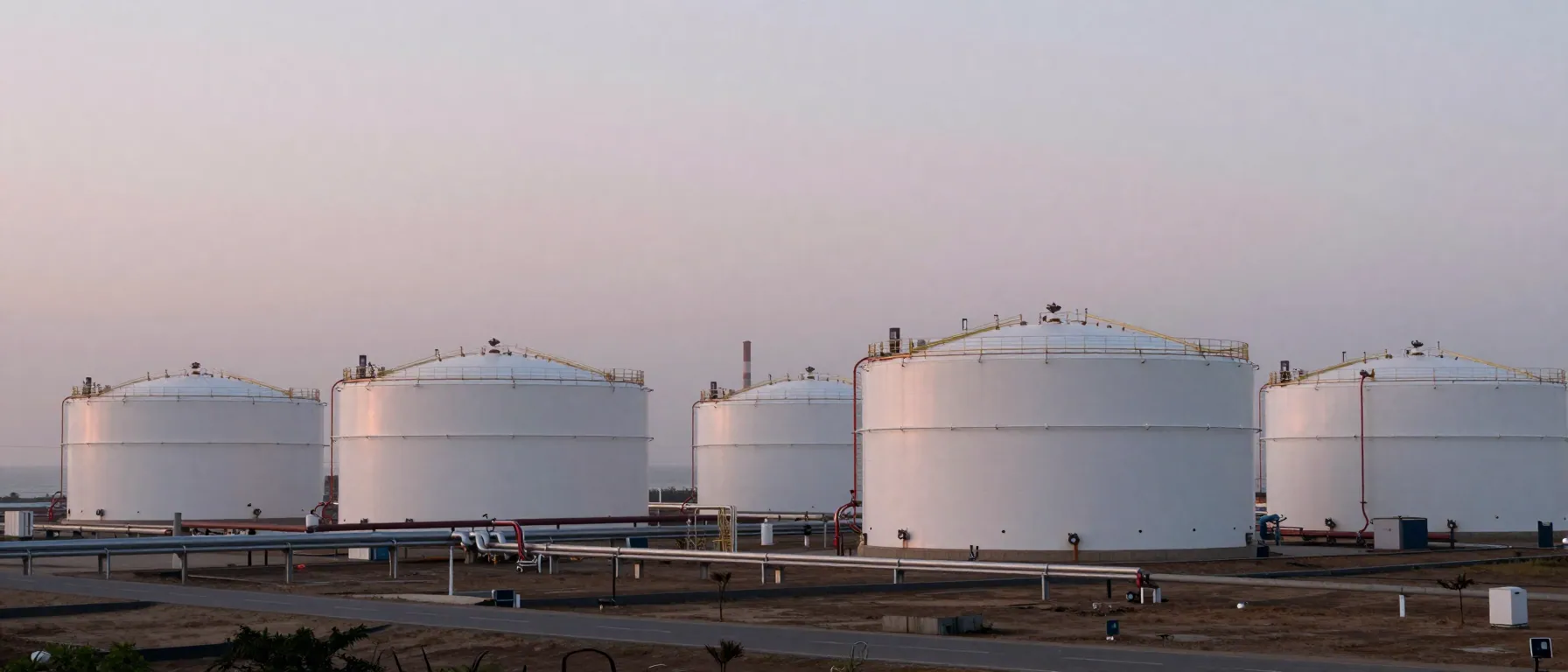 Wide dawn view of white fuel storage tanks and pipelines across a misty coastal plain under a pale sky.