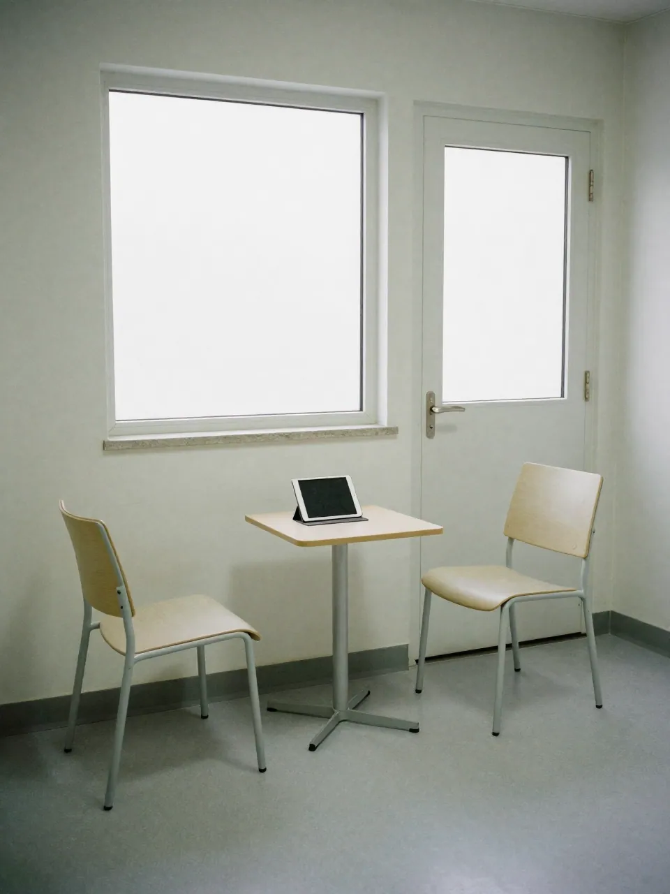 An empty clinical consultation room with two chairs, a side table and a face-down tablet in daylight.