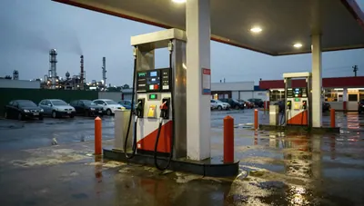 Diesel nozzle in a holster on a filling station forecourt at dusk, wet pavement reflecting canopy lights, refinery skyline behind.