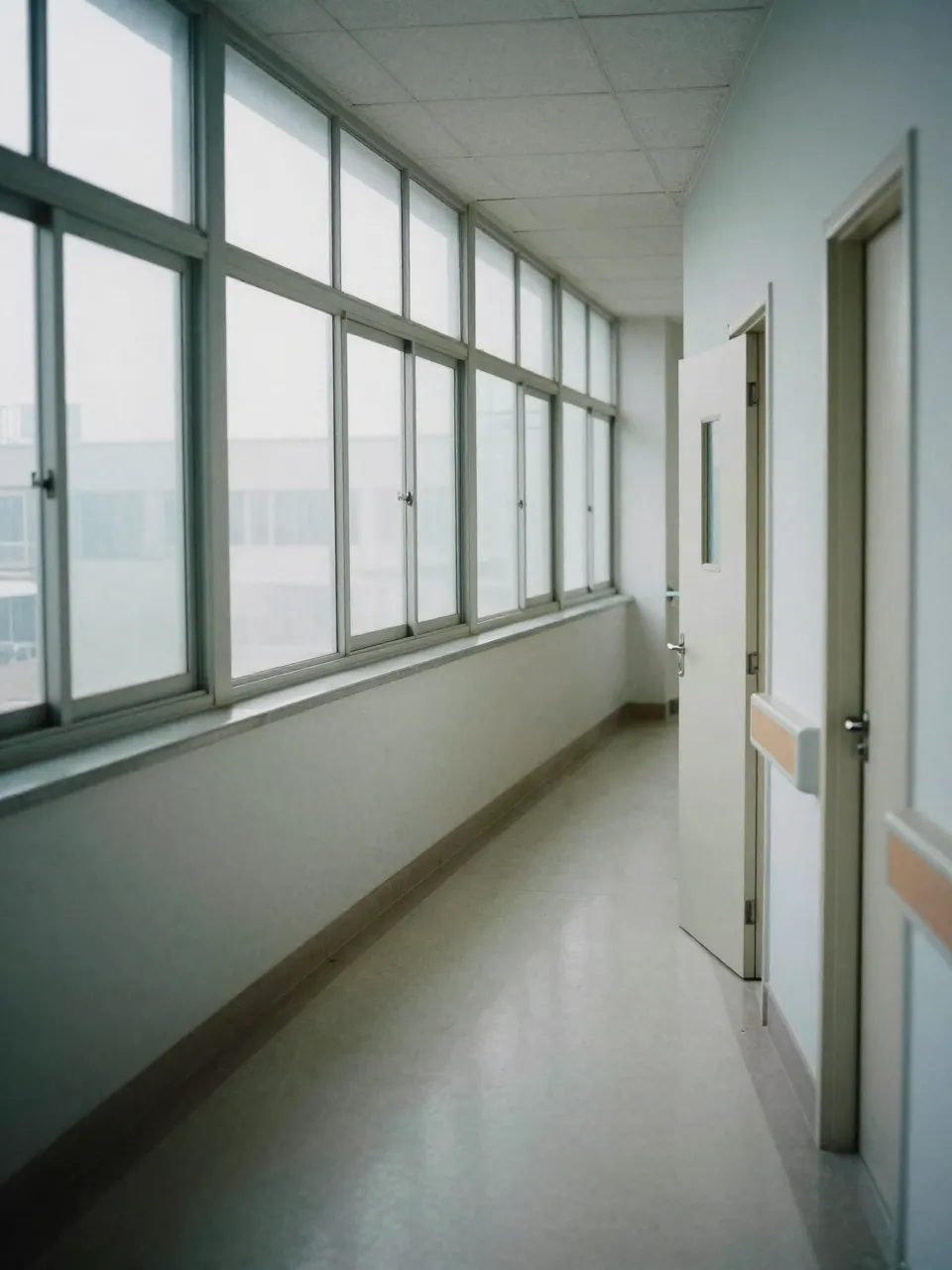 Empty hospital corridor with daylight through tall windows and closed doors.