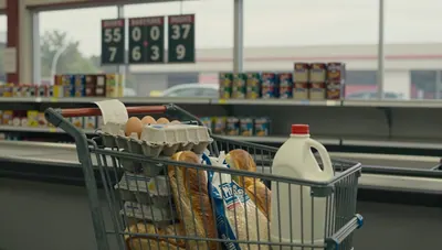 Shopping cart of groceries and a receipt in a supermarket aisle, with a blurred fuel price board outside the window.