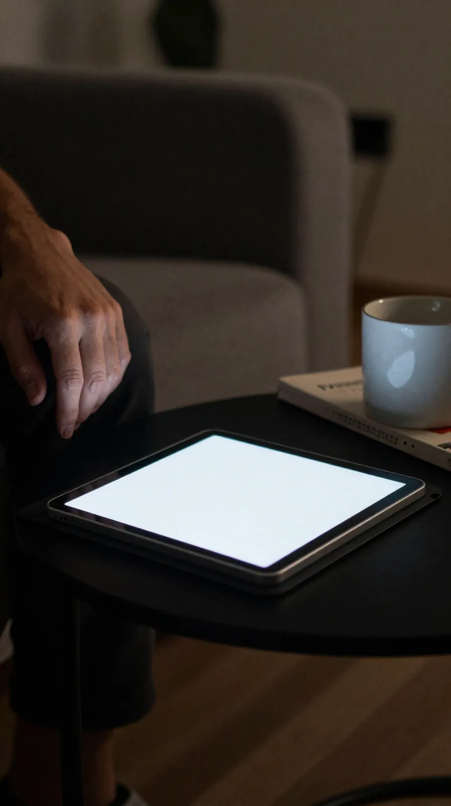 A tablet lies face-down on a side table with faint glow at its edges, a hand withdrawn, quiet living room.