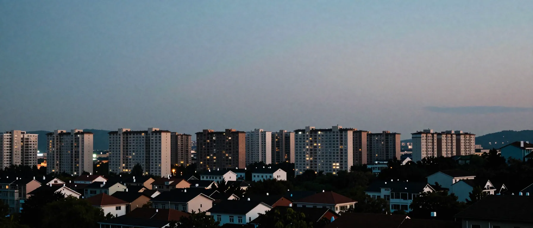 A wide dusk view of a city skyline with apartment blocks and houses from a low hill.