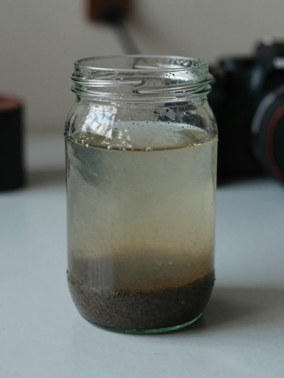A glass jar of cloudy dark fuel with sediment and a watery layer at the bottom.