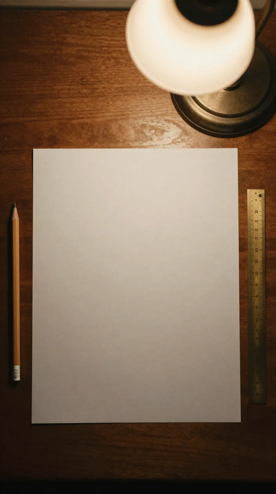 An overhead view of a blank sheet of paper, pencil and brass ruler on a wooden desk.