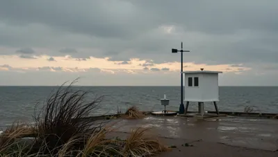 Coastal weather station with instrument shelter, anemometer and rain gauge on a bluff above a calm winter ocean at dusk.